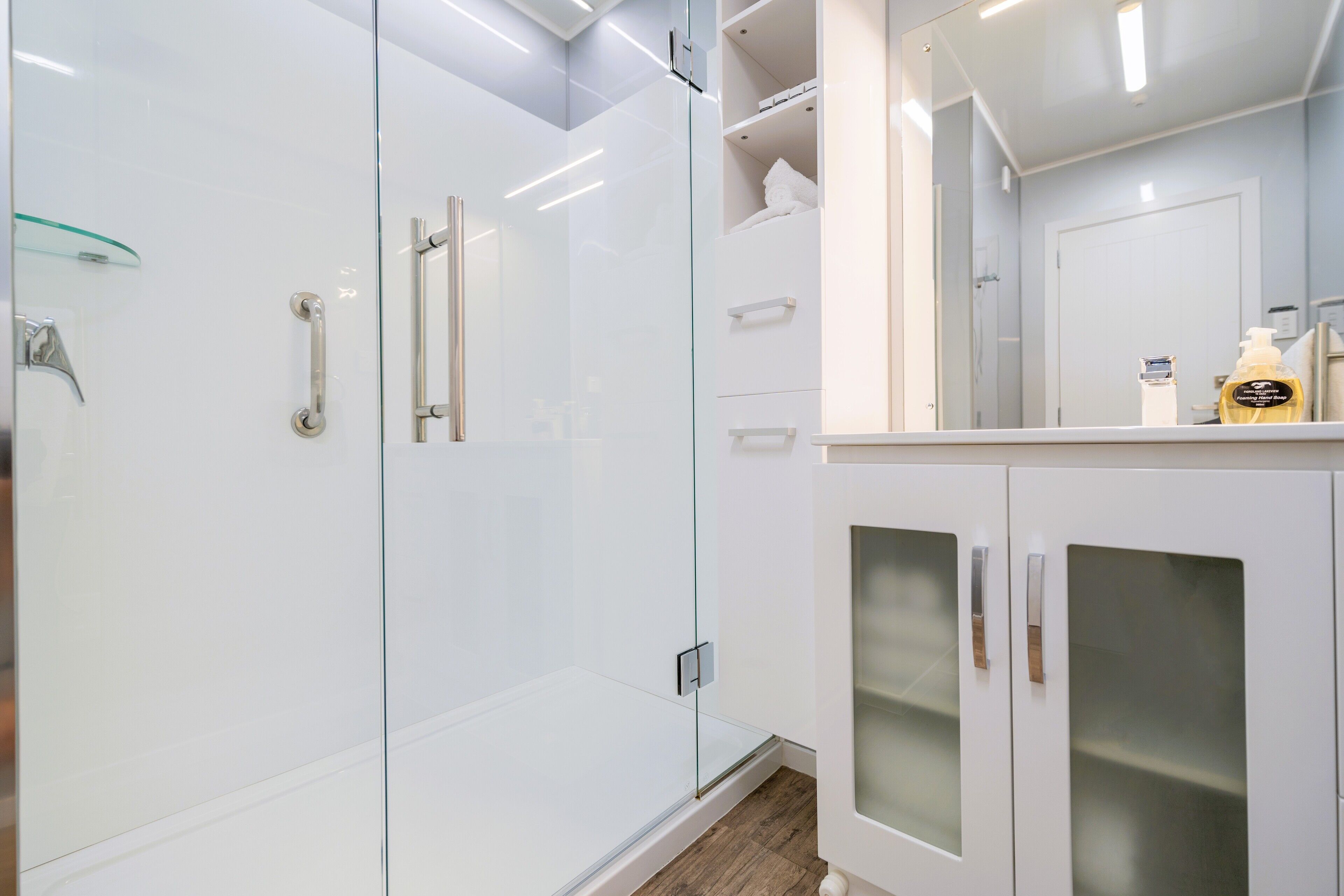 Un baño moderno con cabina de ducha de vidrio, muebles blancos y un espejo que refleja la habitación.