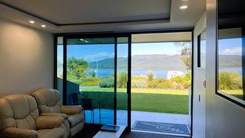 A modern room with a leather sofa, a small table, and a chair. It features large sliding glass doors leading to a balcony with a view of a lake and mountains.