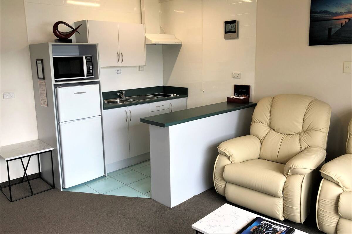 Een moderne keuken met witte kasten, een magnetron en een spoelbak. Er staat een beige fauteuil en een klein tafeltje met een tijdschrift.