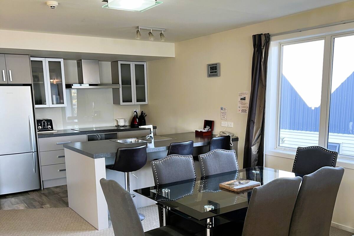 Een moderne keuken met een eethoek, met een tafel met glazen blad, grijze bekleden stoelen en een groot raam met uitzicht.