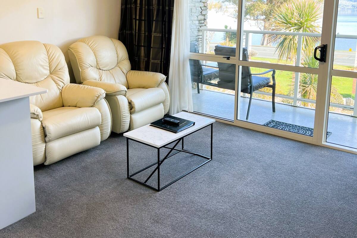 Een woonkamer met een beige bank, een bijzettafeltje en uitzicht op een balkon met buitenmeubilair.