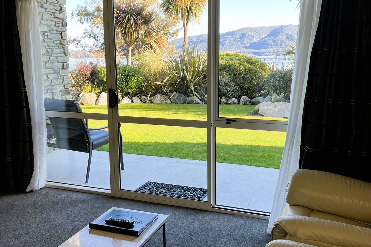 Een kamer met een schuifglazen deur naar een terras met een gazon en uitzicht op bergen. Er staat een witte tafel en een beige stoel in de kamer.