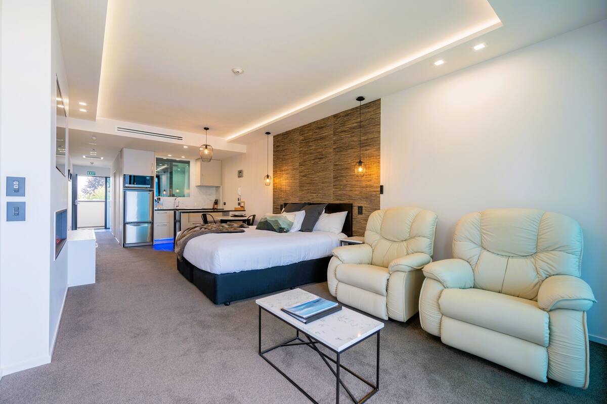 Une chambre d’hôtel moderne avec un lit, deux fauteuils inclinables, une petite table et, au fond, un coin cuisine.