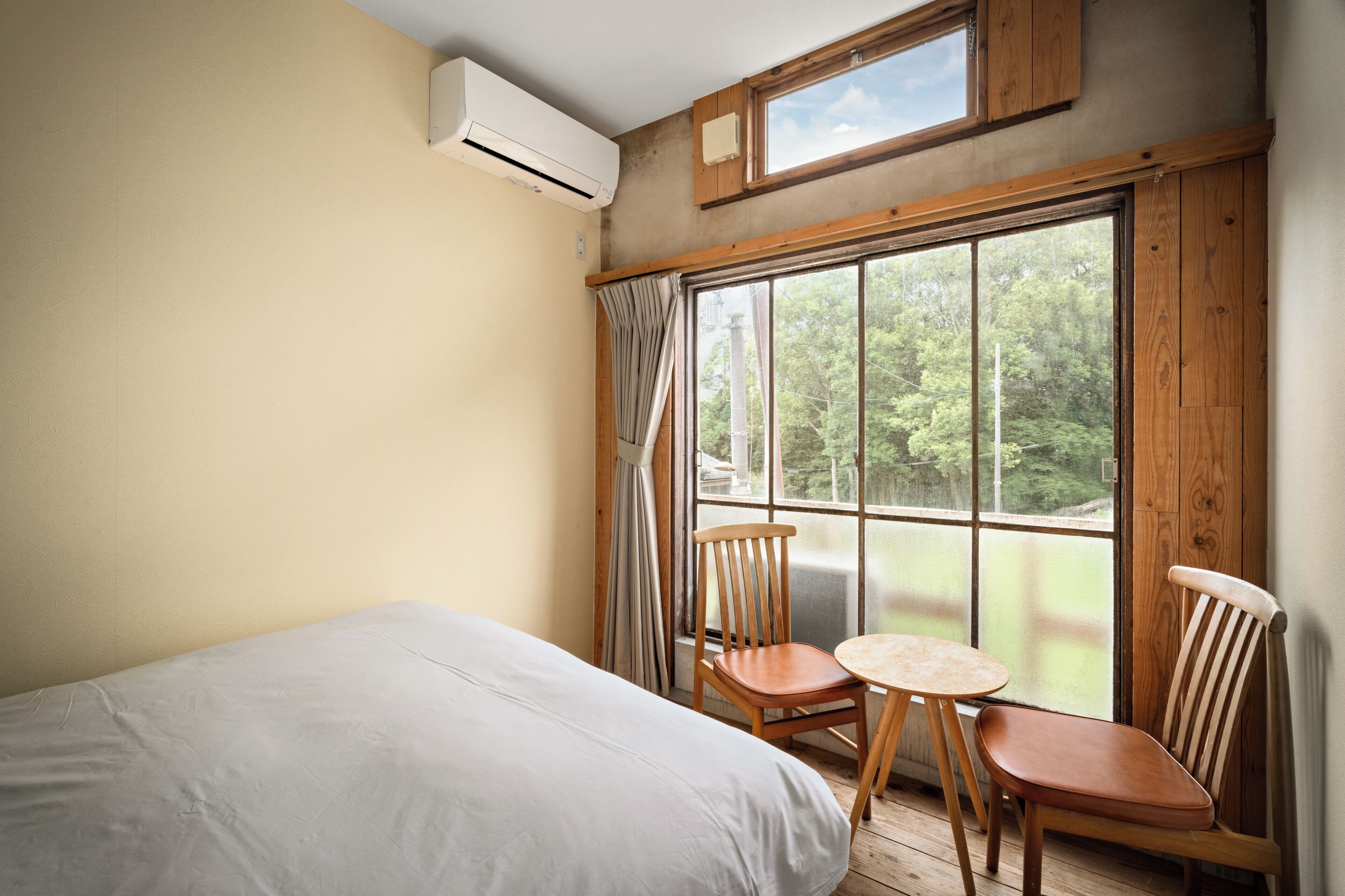 A room with a bed, two wooden chairs, a small table, a large window with curtains, and an air conditioning unit.
