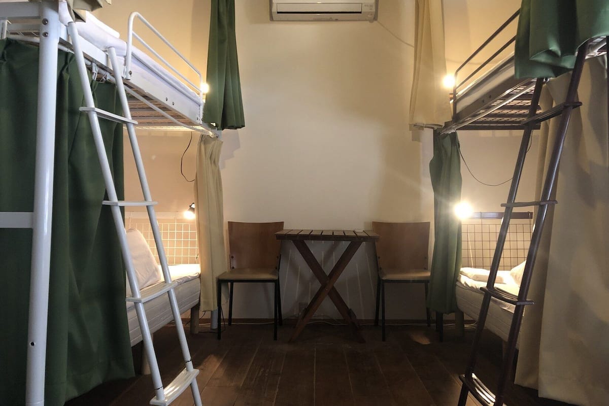 A dormitory room with bunk beds, a small table, two chairs, an air conditioning unit, and a window with curtains.