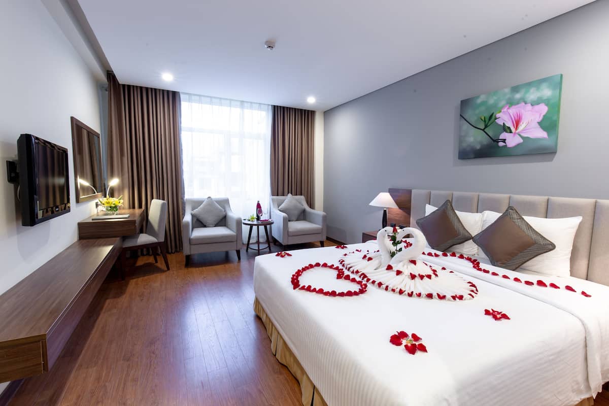 Una habitación de hotel con una cama decorada con pétalos de rosa, un escritorio con lámpara, dos sillones y un cuadro en la pared.