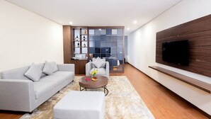 Een moderne woonkamer met een grijze bank, een witte poef, een ronde salontafel met een bloevas, een aan een houten muur bevestigde televisie en een inbouwkast met decoratieve objecten.