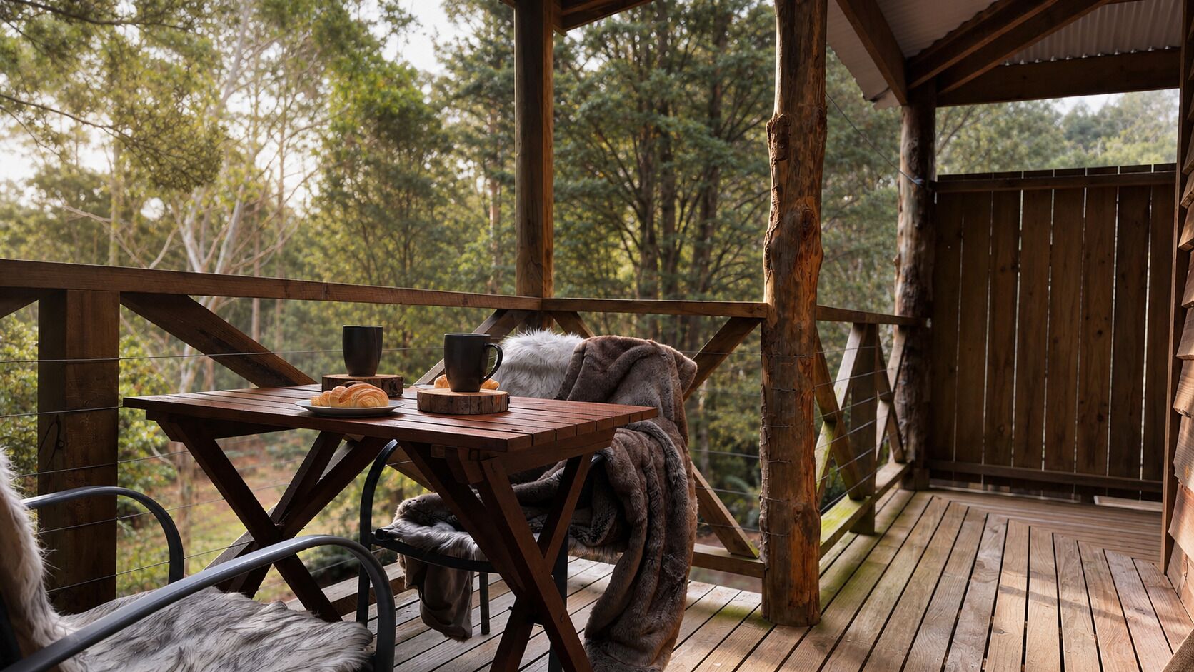 Une terrasse en bois avec une table et des chaises, une couverture et une tasse, le tout entouré d’arbres.