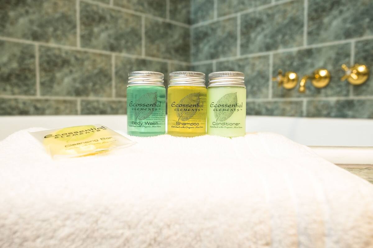 Three bottles of Ecessential Elements body wash, shampoo, and conditioner on a white towel in a bathroom with a tiled wall.