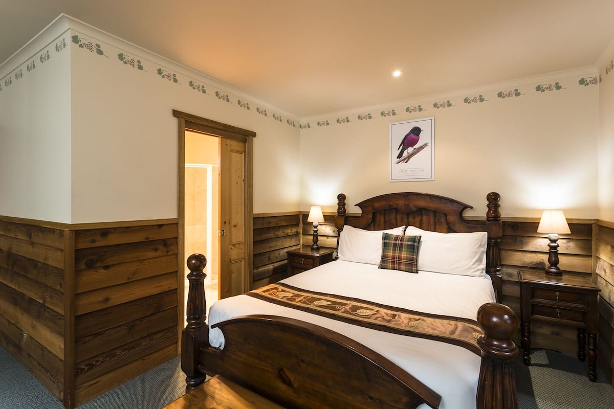 A wooden-paneled bedroom with a large bed, two bedside tables with lamps, and a door leading to another room.
