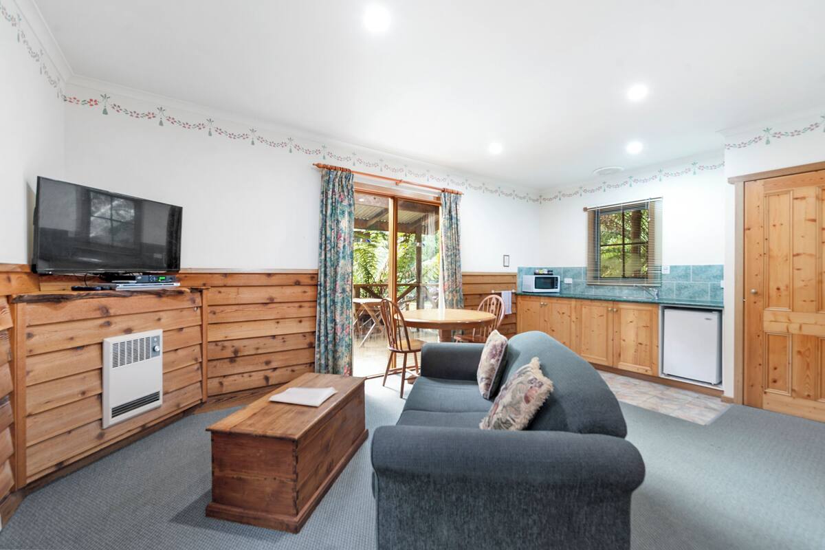 A cozy room with a sofa, wooden walls, a television, a small table, and a kitchen area with a refrigerator and microwave.