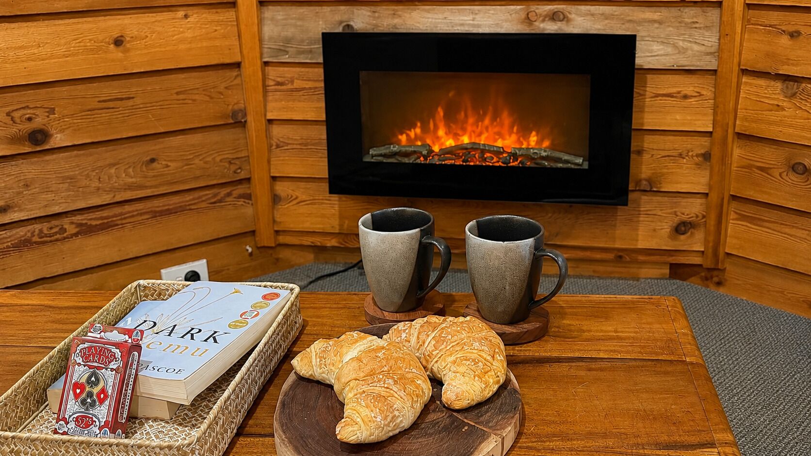 A cozy room with a fireplace, a table with a tray of playing cards, two mugs, and a plate of croissants.