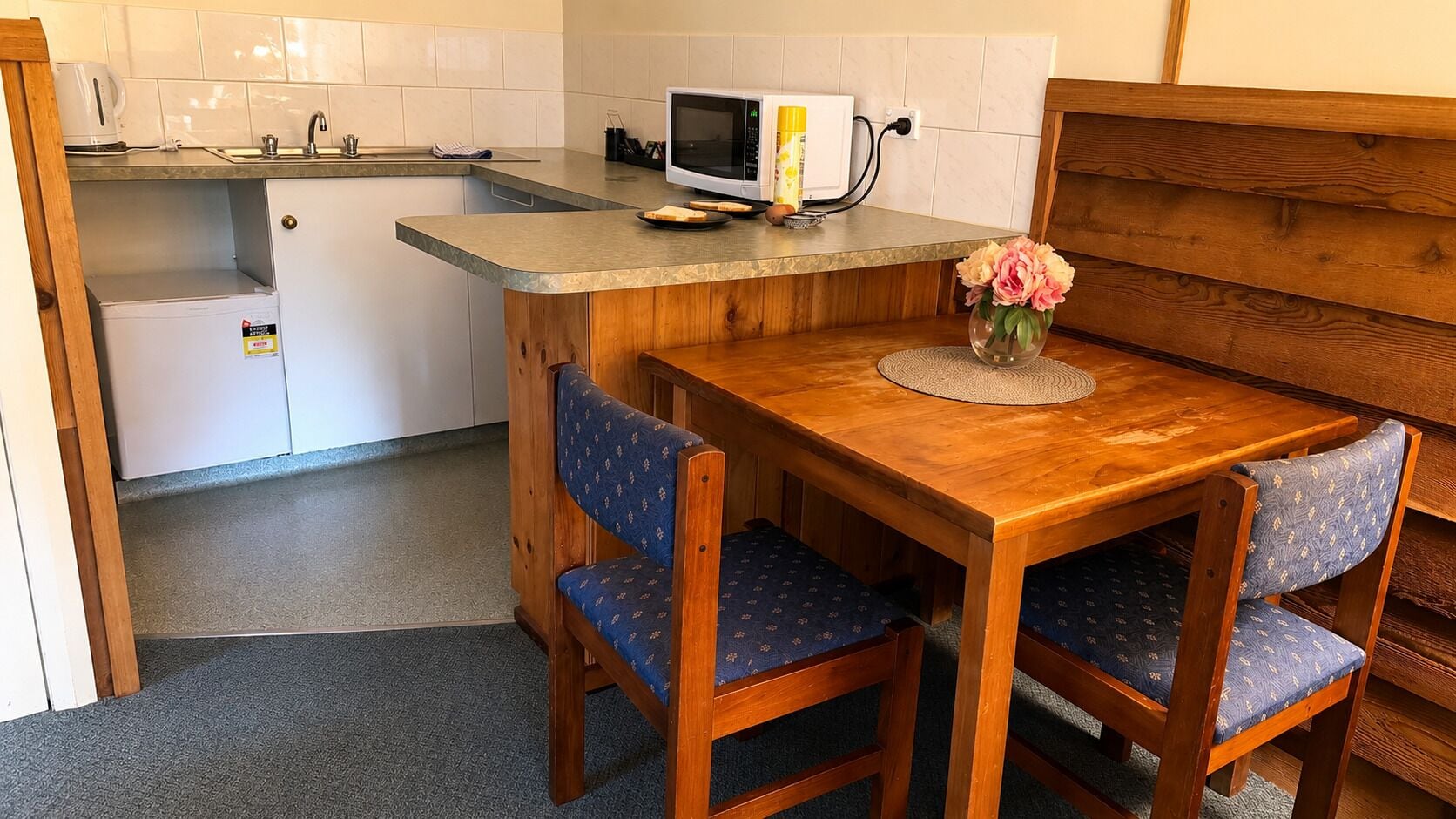 A compact kitchen with a microwave, toaster, and sink. There is a wooden dining table with chairs and a vase with flowers.