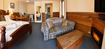 Habitación de hotel con cama, sofá, banca de madera, televisor, chimenea y baño a la vista a través de una puerta abierta.