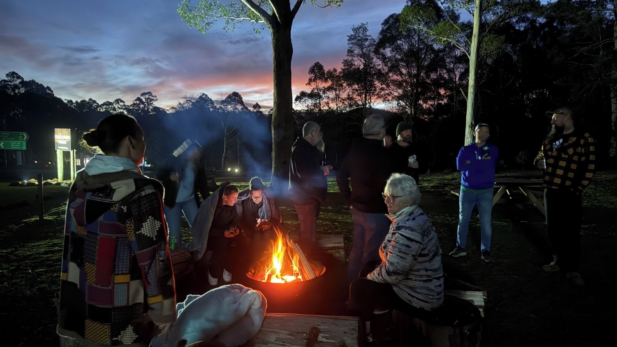 Un groupe de personnes s’est rassemblé autour d’un feu de camp dans un cadre extérieur, avec des arbres et une montagne en arrière-plan.
