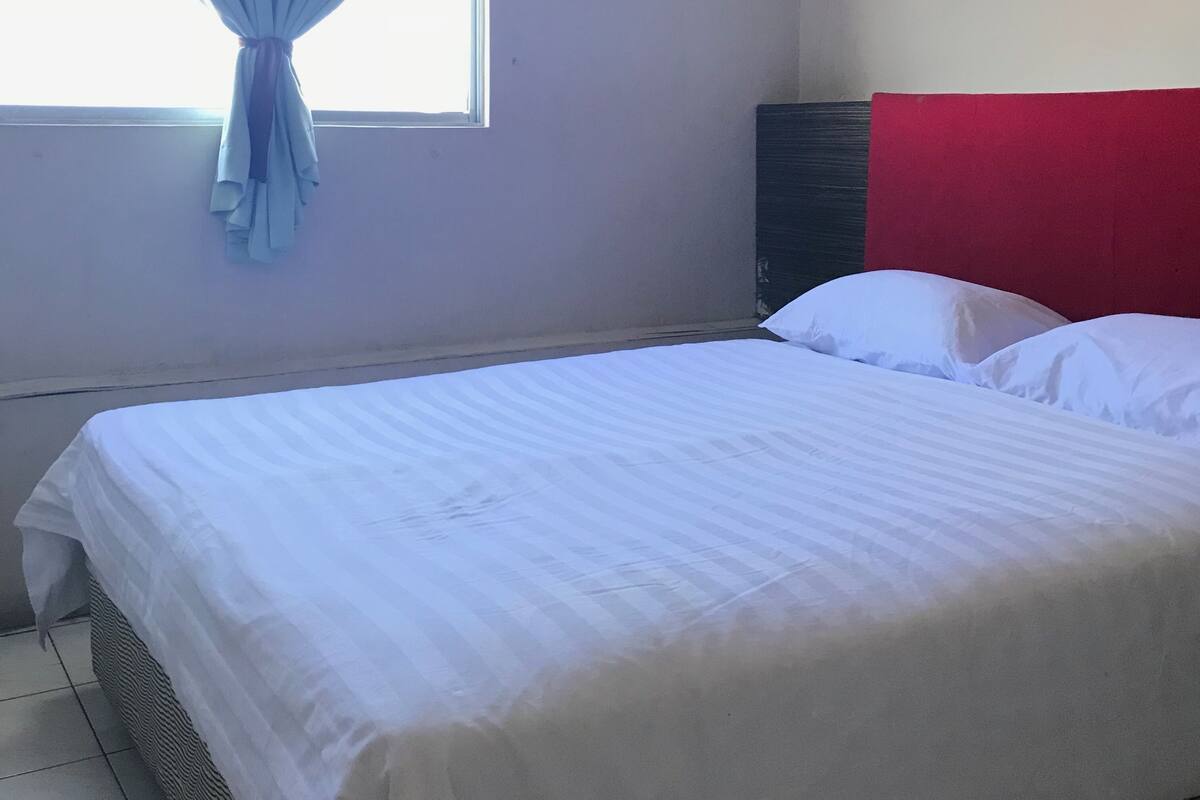 Een enkel bed in een kamer met raam, een rood hoofdeinde en een gordijn.