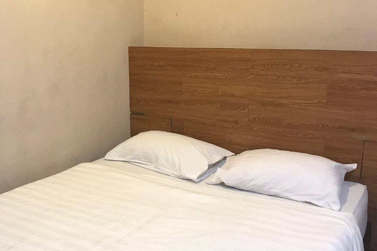 Een enkel bed met wit beddengoed en kussens, een houten hoofdbord en een effen muur.