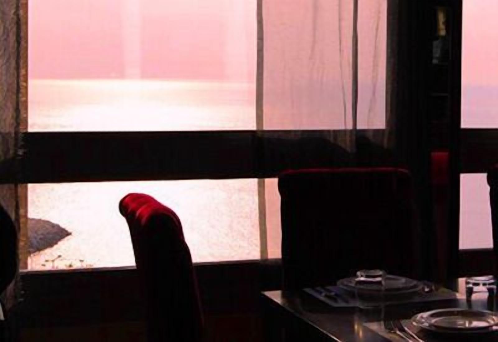 Ein Zimmer mit großem Fenster und Blick auf ein Gewässer. Ein Stuhl und ein Tisch mit Teller und Glas sind vorhanden.