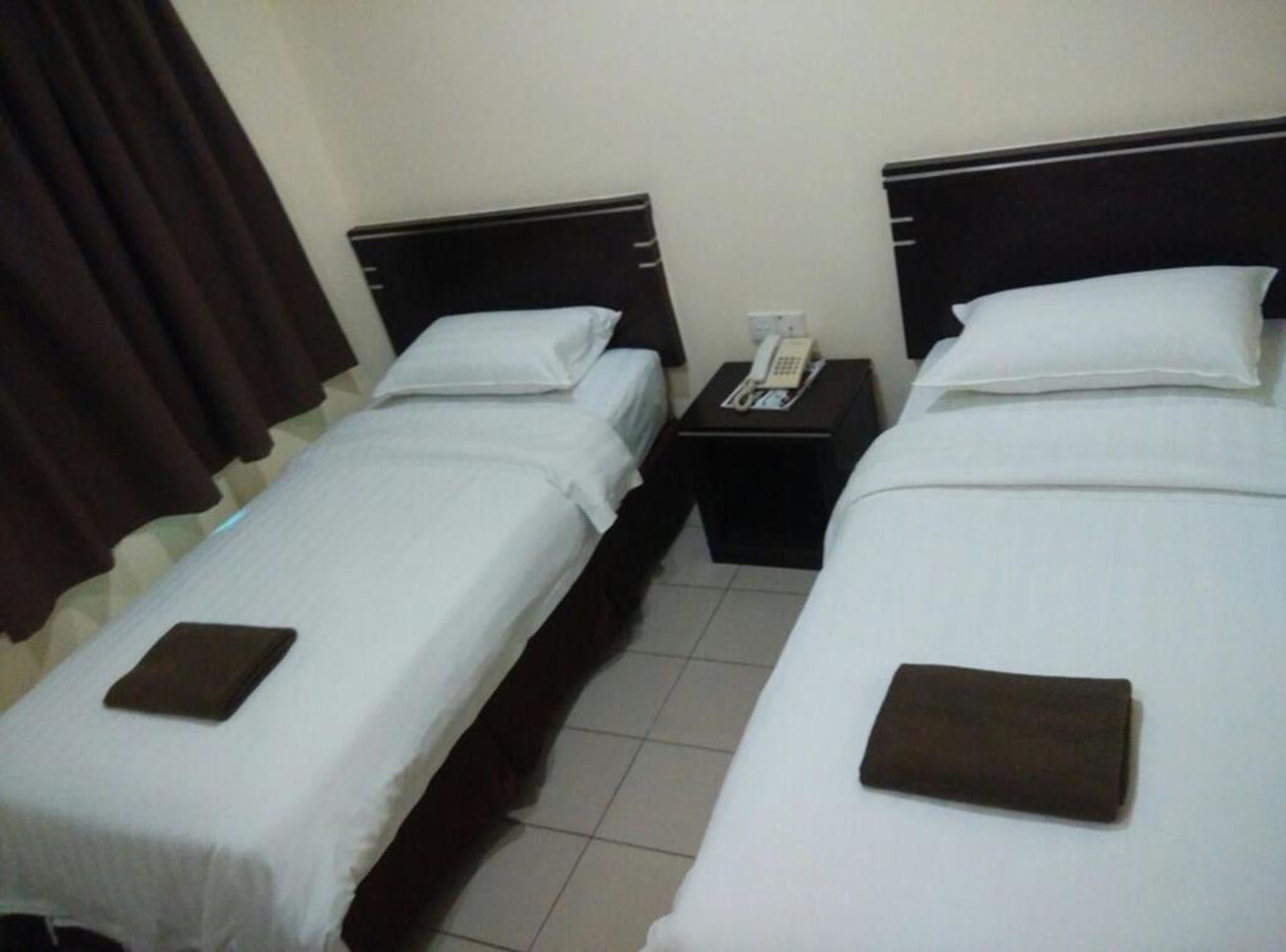 Habitación de hotel con dos camas individuales, cada una con una almohada y un objeto marrón sobre la cama. Hay una mesita de noche entre las camas con un teléfono encima.