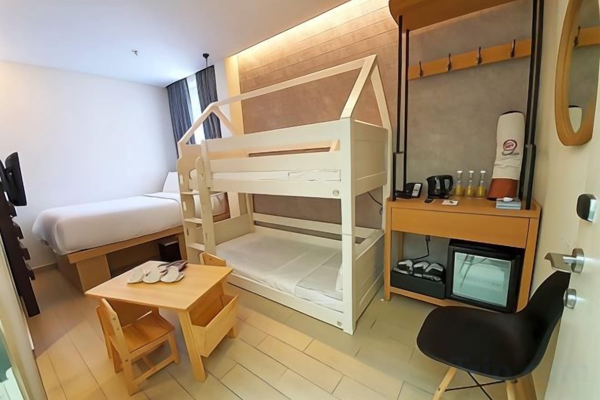 A room with a bunk bed, a desk, a chair, a small table, a mirror, and a shelf with toiletries.
