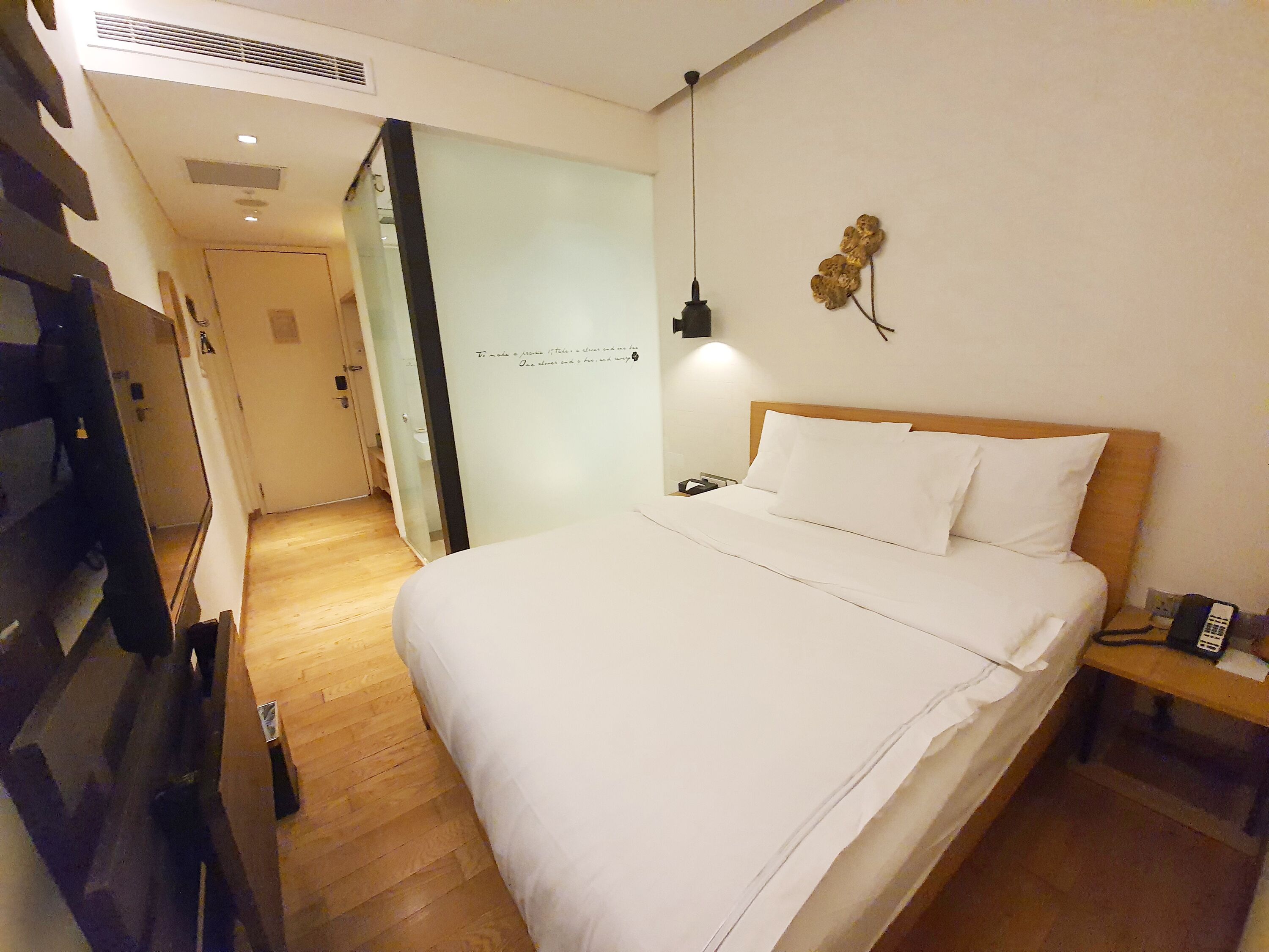 Habitación de hotel con piso de madera, una cama con sábanas blancas, mesita de noche con teléfono y lámpara de pared.