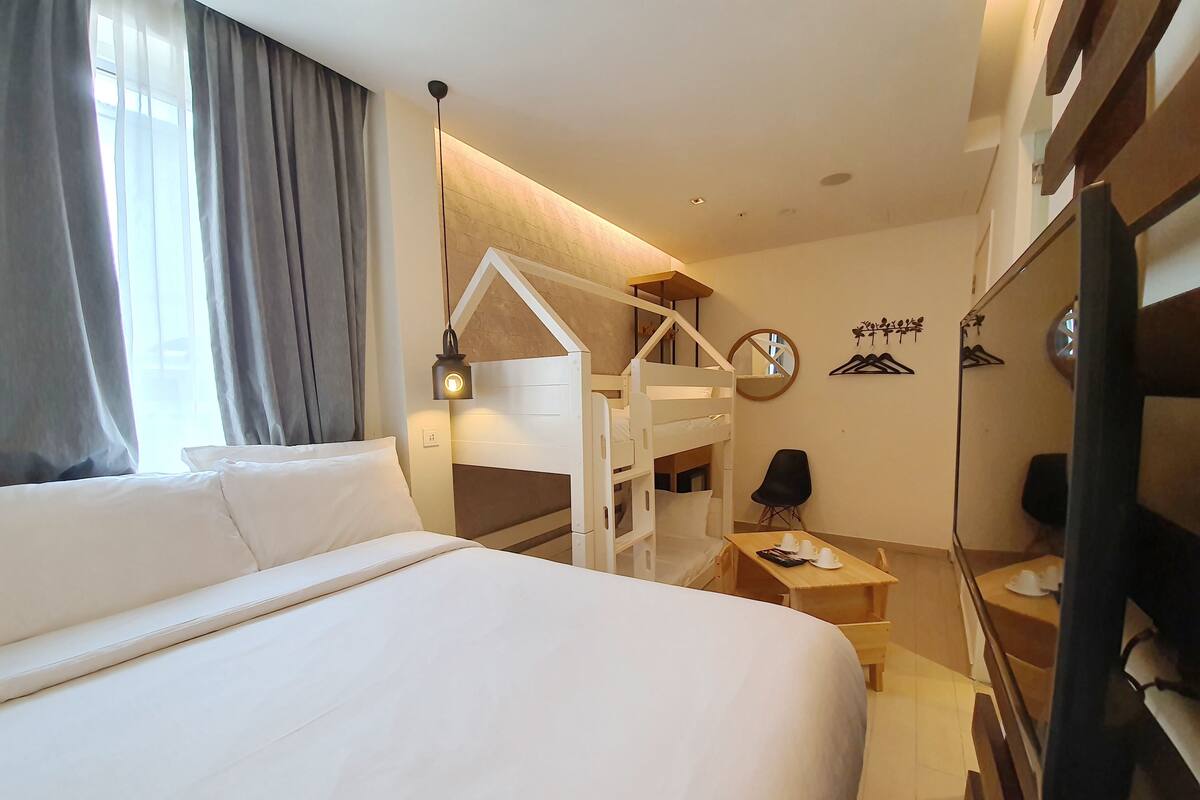 A hotel room with a bunk bed, a single bed, a small table with cups and a tray, a chair, a mirror, and a coat rack.