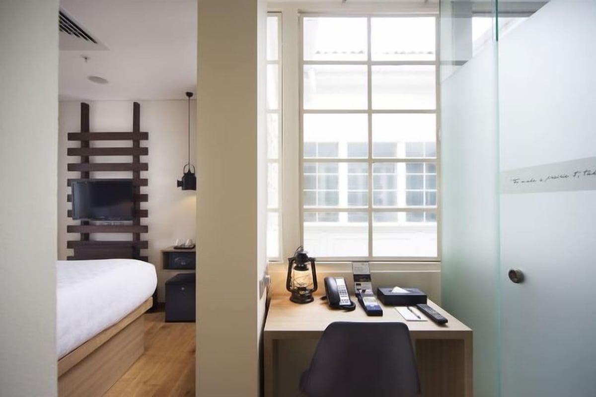 Quarto de hotel pequeno, com uma cama, uma mesa com computador, uma luminária e uma janela com vista.