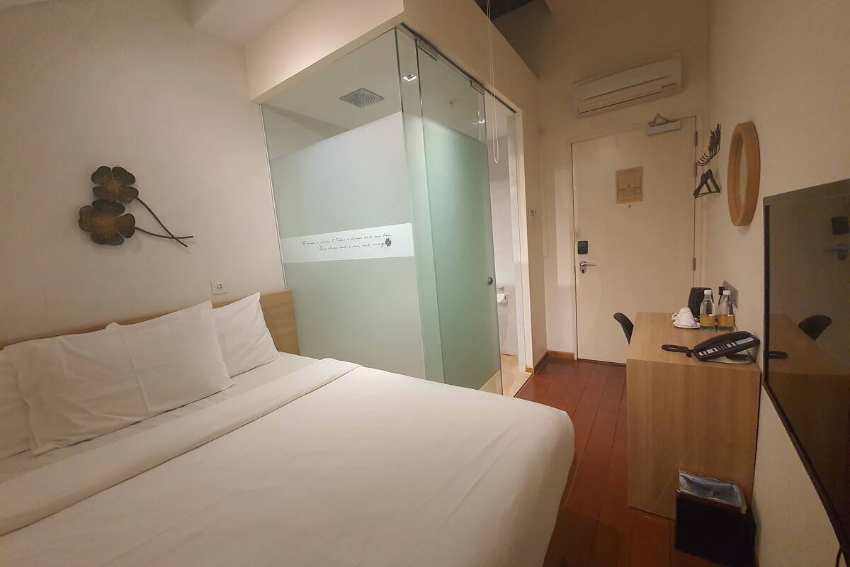 A hotel room with a bed, a desk with a phone, a mirror, and a small trash bin.