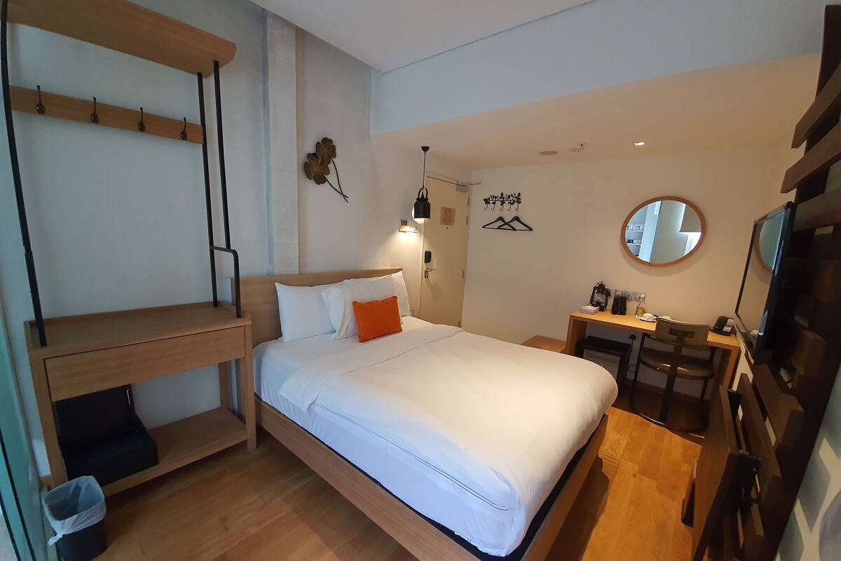 A modern hotel room with a wooden bed, a desk with a chair, a small table, and a mirror.