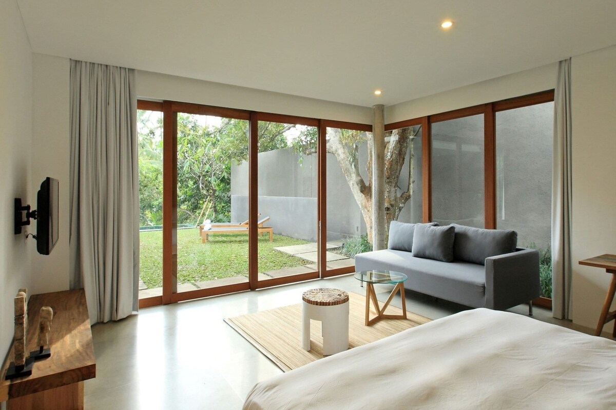 A modern bedroom with a large bed, a sofa, a glass table, and a TV mounted on the wall.