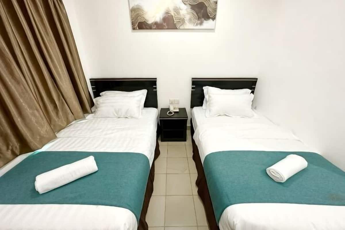 A room with two single beds, each with a pillow and a rolled towel. There is a nightstand between the beds with a lamp.
