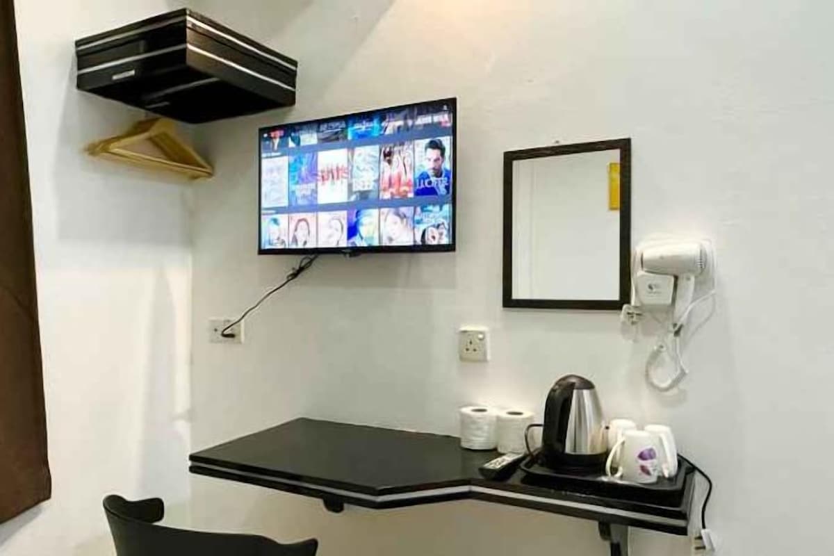 Une chambre d’hôtel équipée d’un téléviseur à écran plat fixé au mur, diffusant différentes chaînes, d’une petite table avec une bouilloire et des tasses, d’un miroir et d’un téléphone.