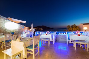 An outdoor dining area with white tables and chairs, a stone wall, and a view of the sea and a mountain at dusk. - Zinbad Hotel (Kas)