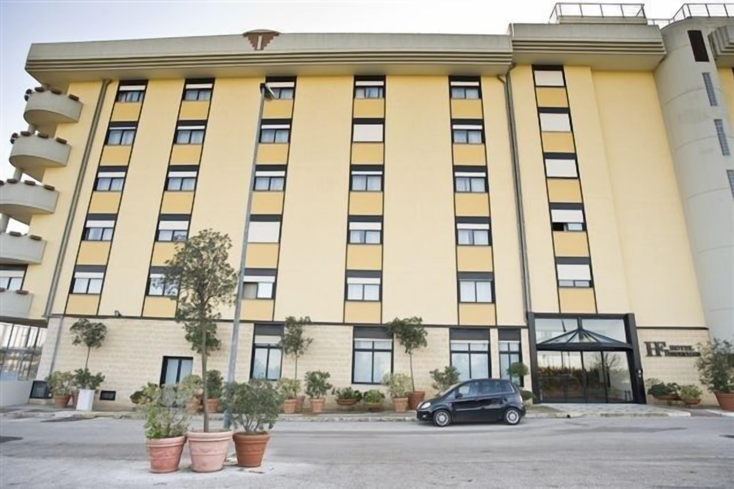 Un hotel de varios pisos con fachada amarilla, múltiples balcones y un estacionamiento con plantas en macetas.