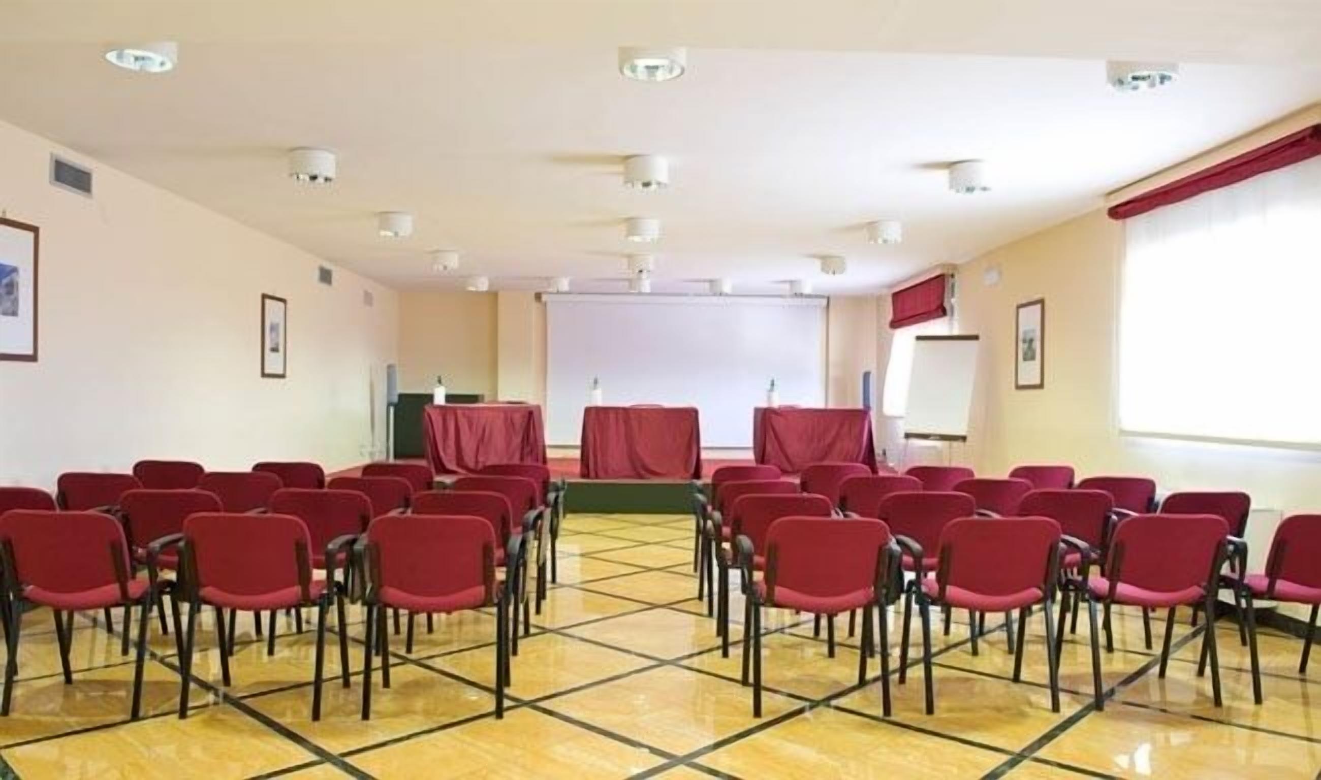 A conference room with rows of red chairs, a stage with a red backdrop, and a projector screen.