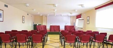 Un salón de conferencias con filas de sillas rojas, un escenario con un fondo rojo y una pantalla de proyector.