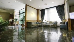 A spacious living room with a large sectional sofa, a coffee table, and a television. The room has a polished floor, large windows with curtains, and several potted plants. - Hotel Federiciano (Valenzano)