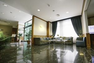 Amplia sala de estar con un sofá modular grande, una mesa de centro y un televisor. El piso es pulido, hay ventanas grandes con cortinas y varias macetas con plantas.