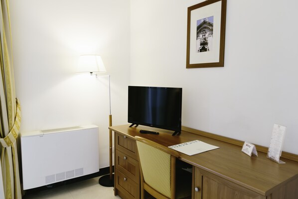 Habitación de hotel con escritorio de madera, televisor de pantalla plana, lámpara de pie, un cuadro en la pared y un radiador.
