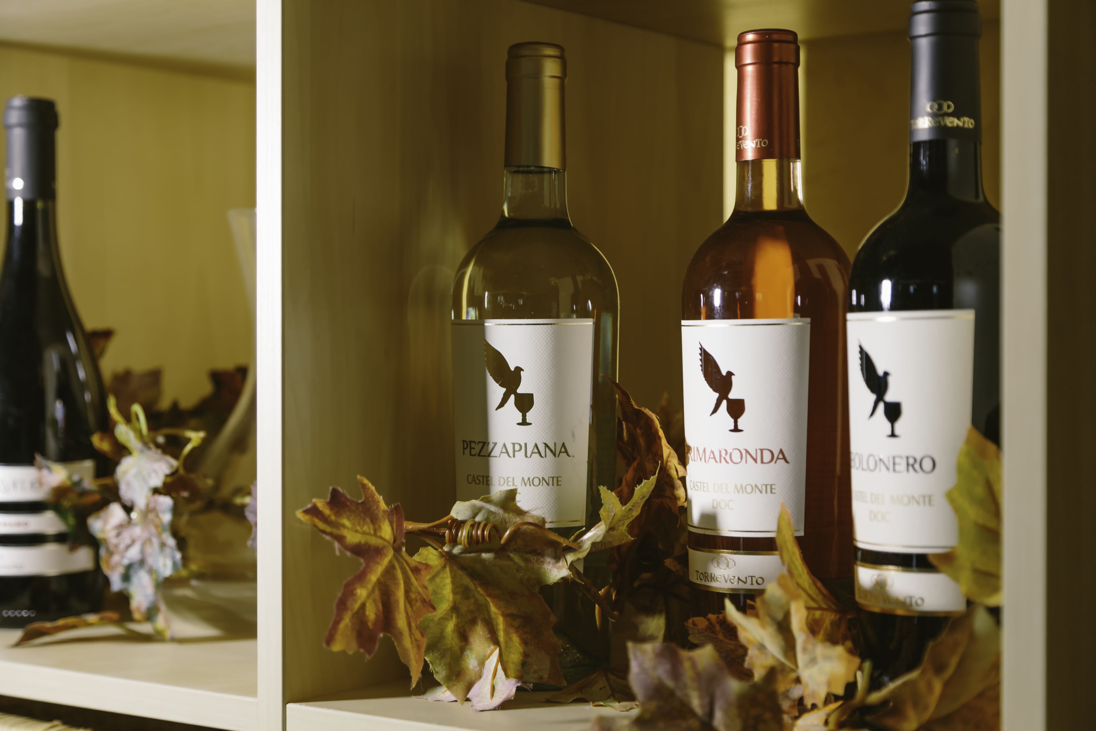 Tres botellas de vino en una estantería con etiquetas y un arreglo decorativo de hojas.