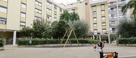 Un terrain de jeux avec des balançoires et un jeu à ressort, situé dans un environnement urbain, entouré d’immeubles à plusieurs étages.