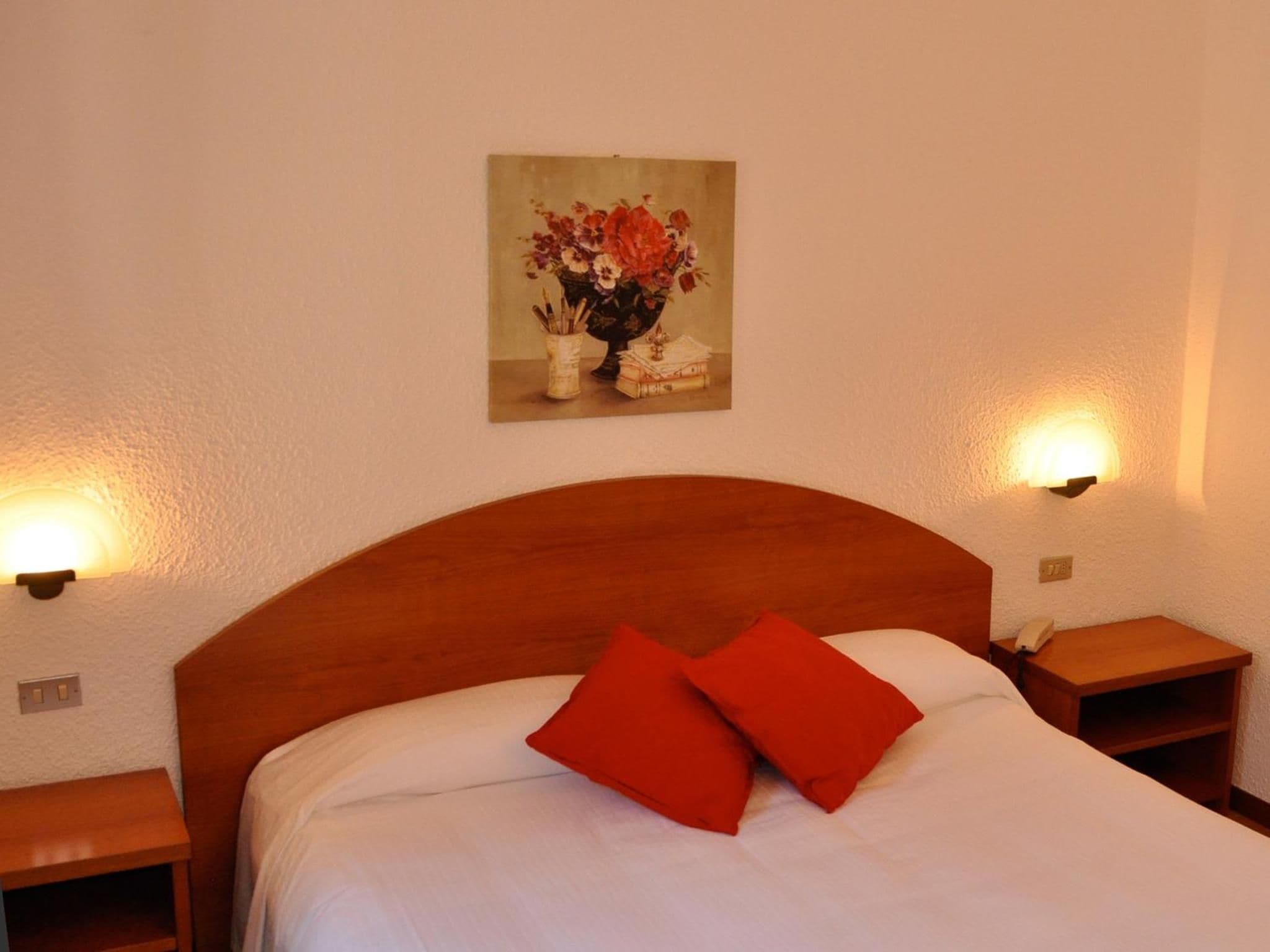 A hotel room with a wooden headboard, two bedside tables, a bed with white bedding and two red pillows, a wall-mounted painting of flowers, and two wall-mounted lamps.