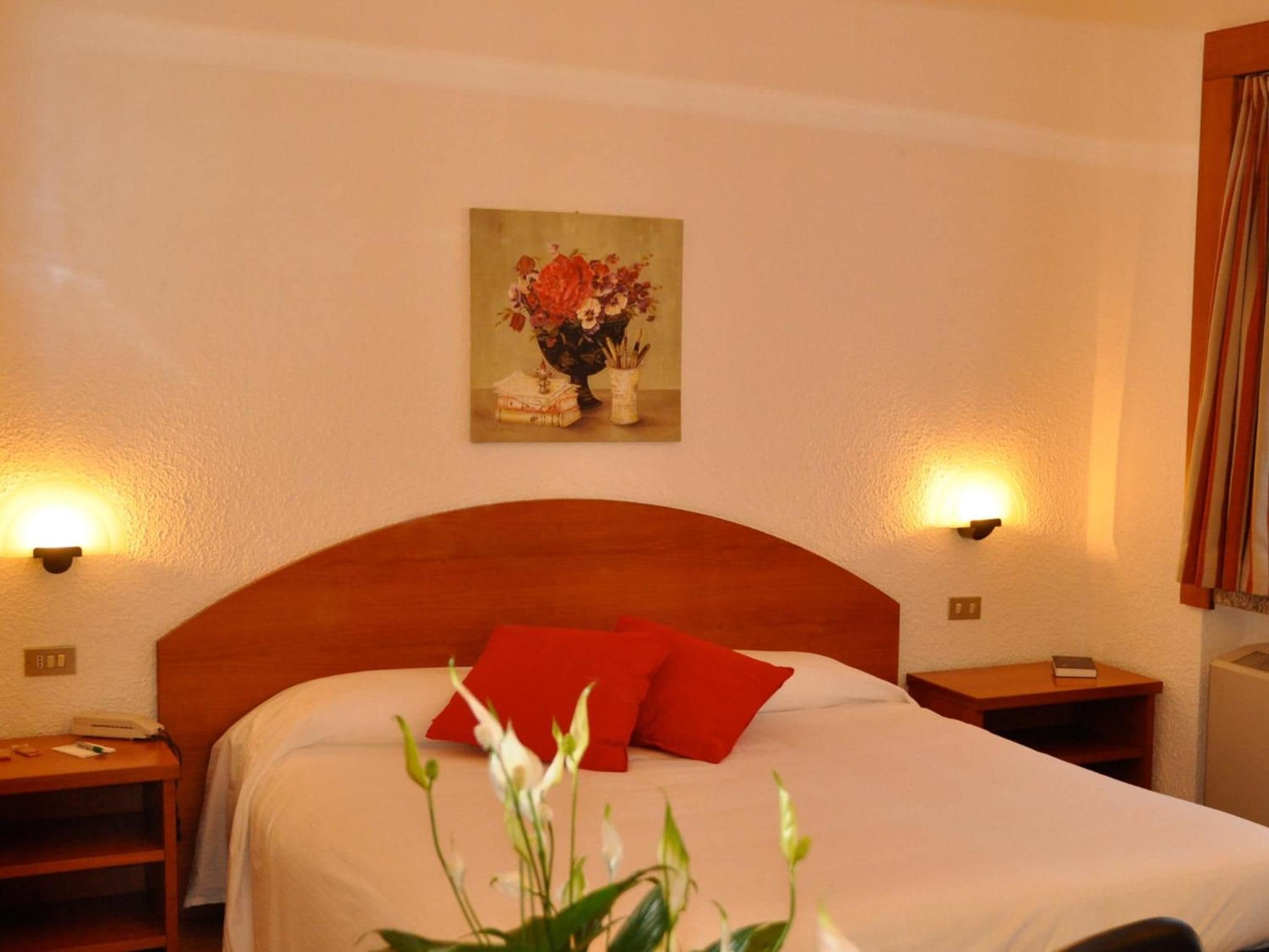 A hotel room with a wooden headboard, a bed with white bedding and red pillows, a nightstand with a lamp, and a painting of flowers on the wall.