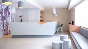 A modern reception area with a white reception desk, a seating area with a sofa and chairs, and a large window providing natural light. - The Grey Hotel (Dortmund)