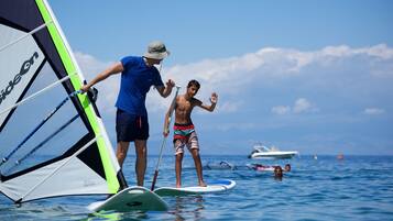 A person on a paddleboard with a sail, another person standing nearby, and a boat in the background.