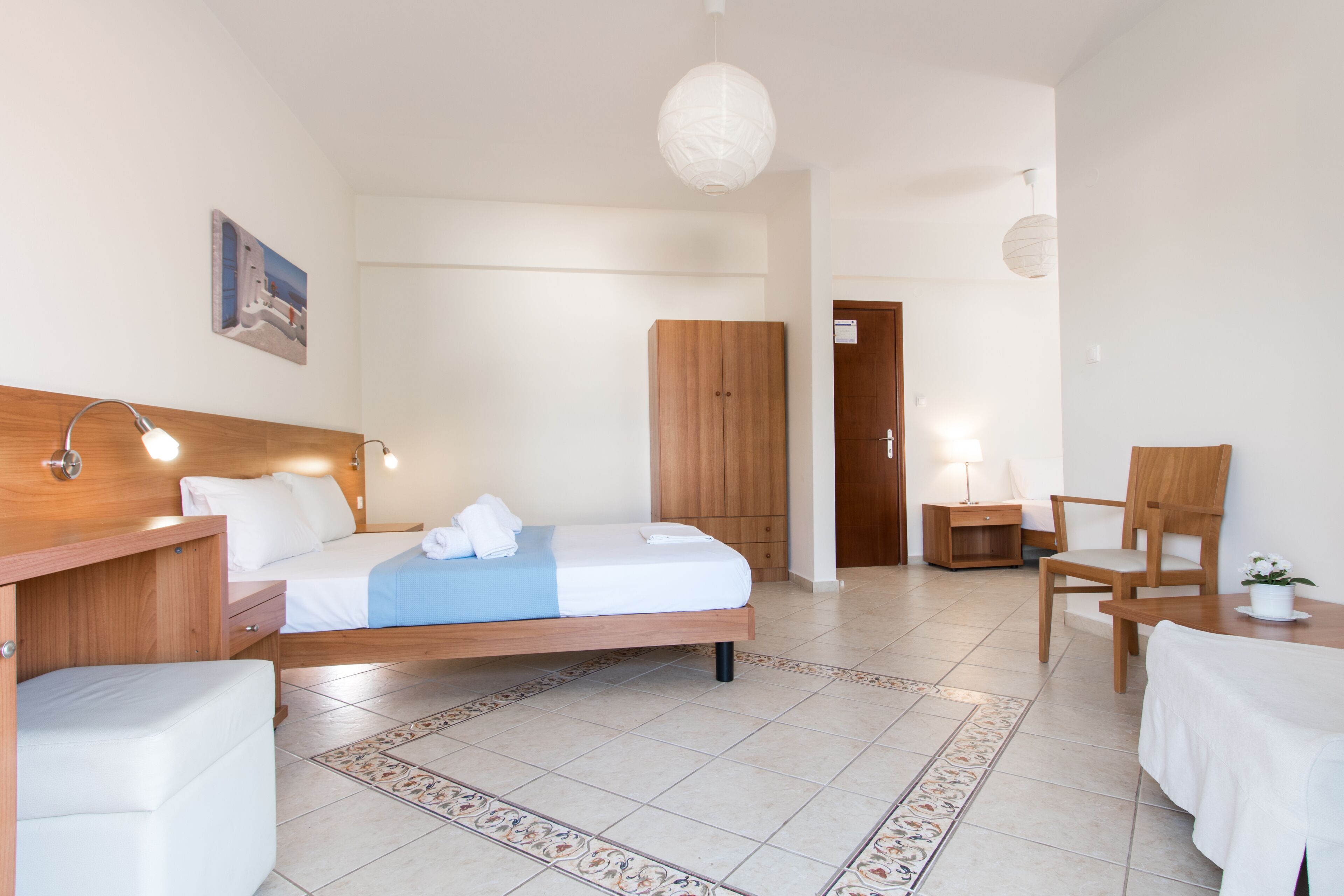 Una habitación de hotel moderna con una cama de madera, un armario de madera, una mesita pequeña con una silla y una mesita de noche con una lámpara.