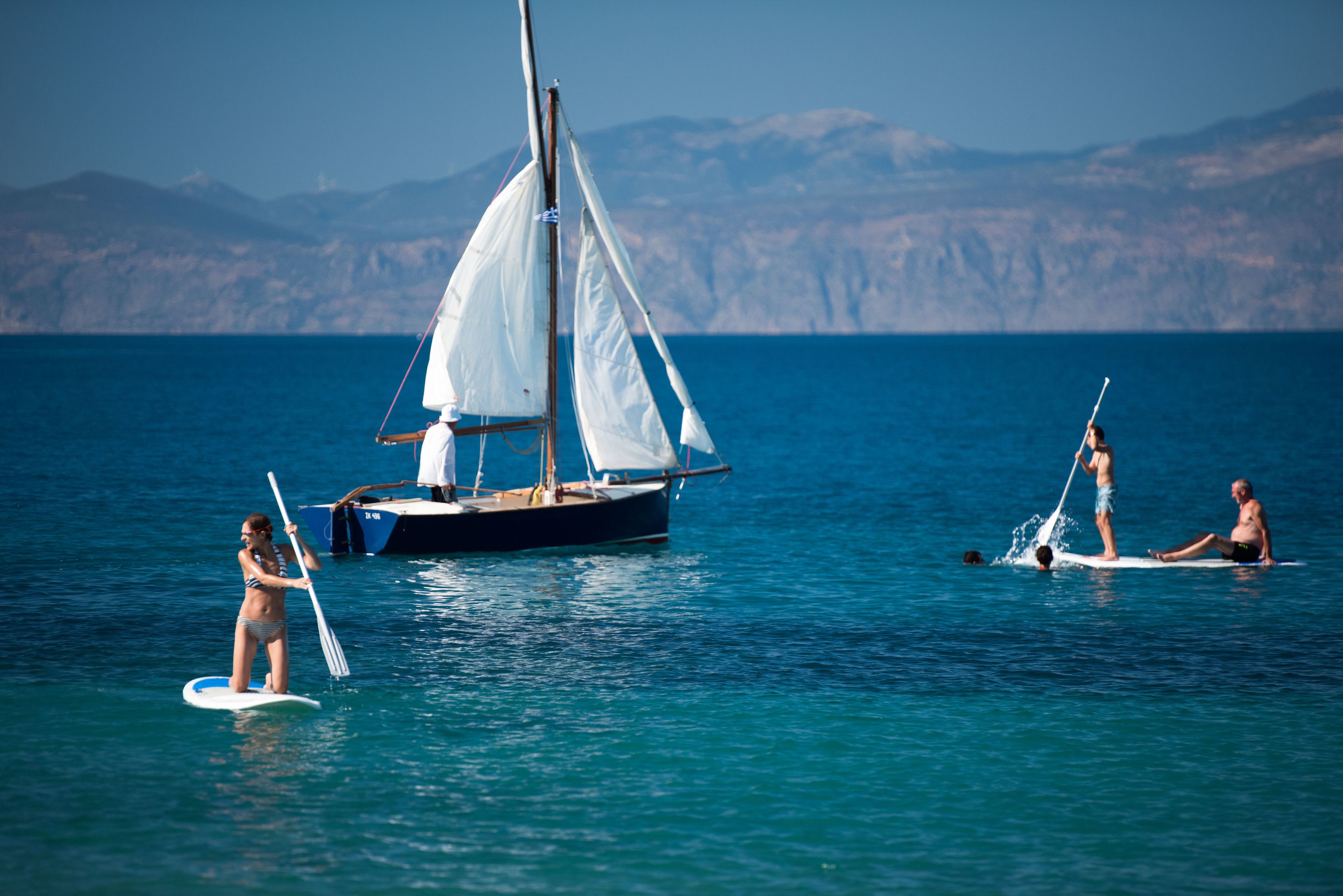 Un velero con gente en tablas de remo en el agua, cielo despejado y montañas a lo lejos.