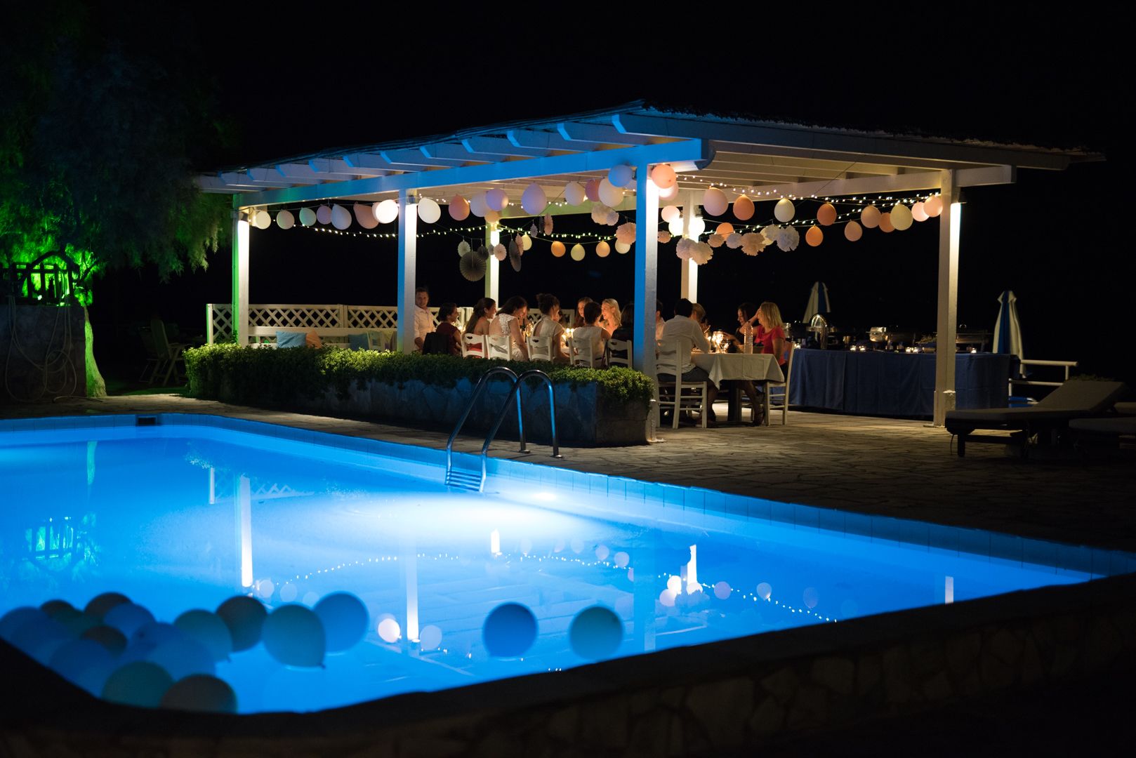 A poolside area with a covered seating area and tables, surrounded by string lights and decorative elements.