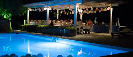 A poolside area with a covered seating area and tables, surrounded by string lights and decorative elements.