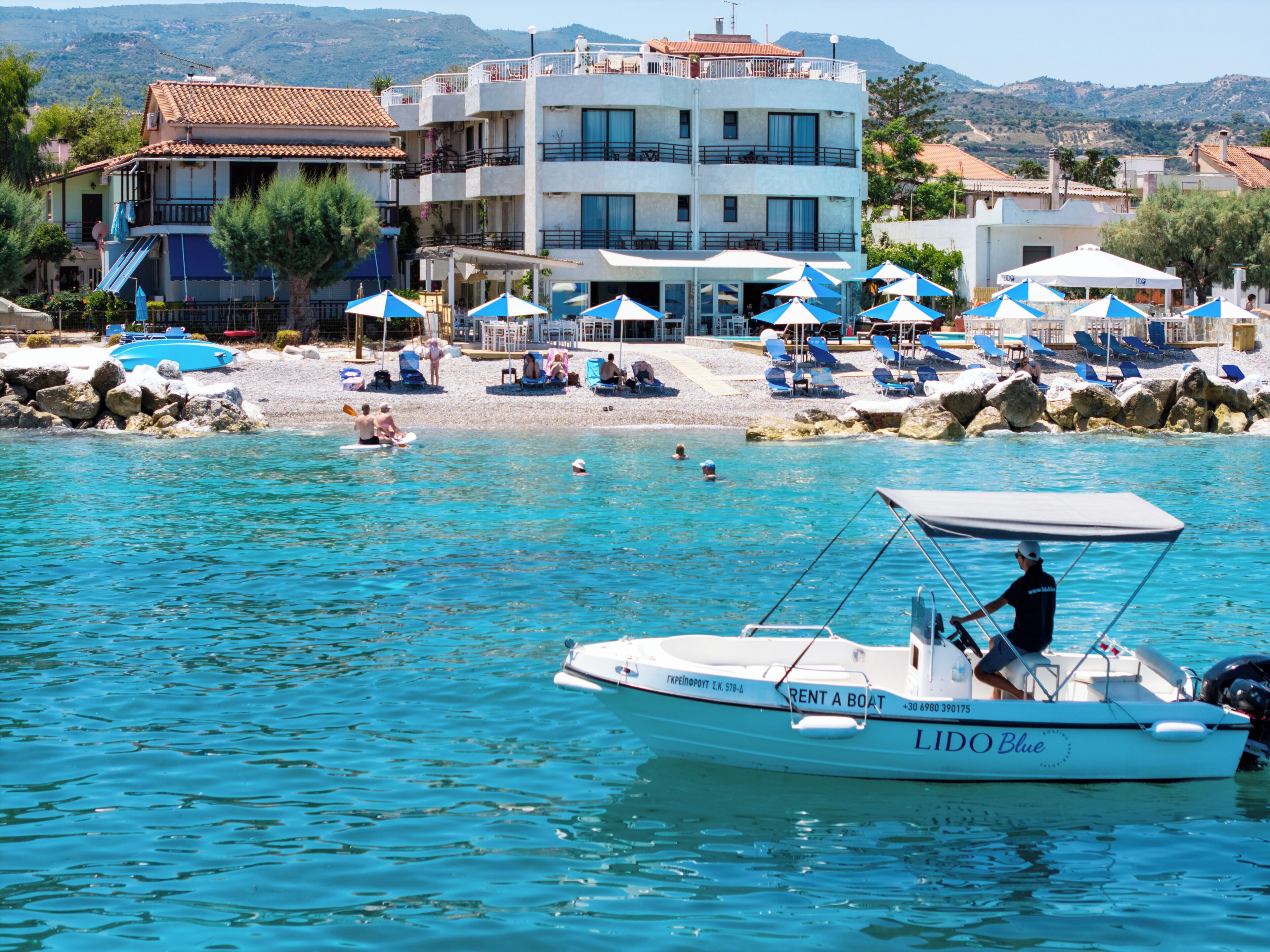 Un service de location de bateaux sur une étendue d’eau calme, avec une plage et des bâtiments en arrière-plan.